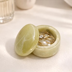 May include: A small, oval-shaped, light green jewelry box with a matching lid. The box is open, revealing gold rings and pearl jewelry inside. The box is made of a stone-like material and sits on a white surface.