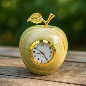 Puede incluir: Un reloj decorativo con forma de manzana, con tallo y hoja dorados. La esfera del reloj es blanca con números y manecillas negras, y un bisel dorado. La manzana es verde y amarilla, y está sobre una superficie de madera.