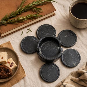 May include: A set of six dark gray marble coasters with a matching holder. The coasters are round with a slightly textured surface. A sprig of rosemary, a cup of coffee, and a plate of snacks are also in the image.
