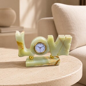 May include: A light green stone desk clock shaped like the word LOVE, featuring a round analog clock face with a gold-tone rim set into the letter O. The piece rests on a rectangular stone base with two small gold-tone spherical supports.