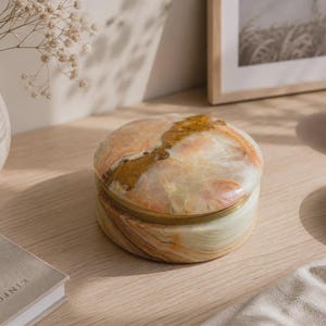 May include: A round stone trinket box featuring a polished surface with swirling patterns of cream, peach, and brown hues. The lid and base are separated by a thin, metallic gold-toned band, creating a smooth, cylindrical silhouette for storage.