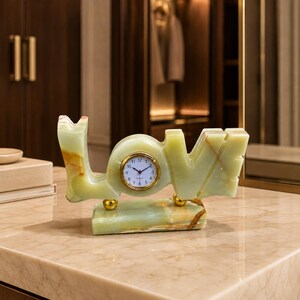 May include: A light green stone desk clock shaped like the word LOVE. The clock face is circular with a gold-tone rim, black numerals, and black hands. The stone features natural veining in shades of cream and amber, resting on a rectangular base.