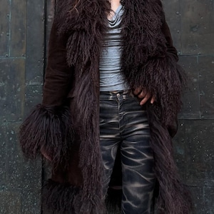 May include: A long, dark brown coat featuring shaggy, fur-like detailing on the collar, cuffs, and front. The coat is paired with a grey top and dark, patterned jeans. The coat's texture and colour give a luxurious, vintage feel.