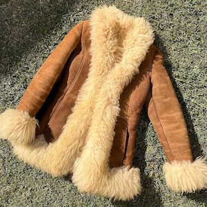 Könnte beinhalten: Braune Wildlederjacke mit cremefarbenem Shearling-Kragen, Manschetten und Besatz. Die Jacke hat ein klassisches Design mit Knopfverschluss und liegt auf grünem Gras. Ein von Vintage inspiriertes Oberbekleidungsstück.