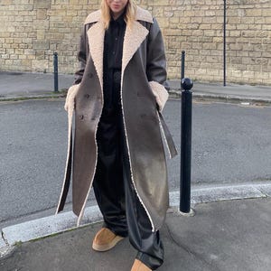 Può includere: Un cappotto lungo marrone con fodera e finiture in montone color crema. Il cappotto ha un design doppiopetto con bottoni grandi e una cintura abbinata. La persona indossa pantaloni neri e scarpe color cuoio.