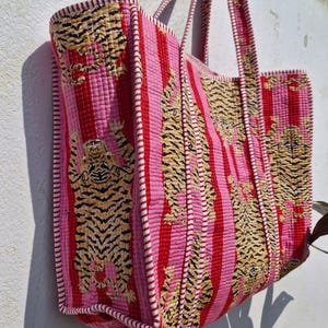 May include: A large tote bag with a quilted design. The bag features vertical stripes in pink and red, with embroidered tiger patterns. The edges are trimmed with white and red stitching, and it has matching handles.
