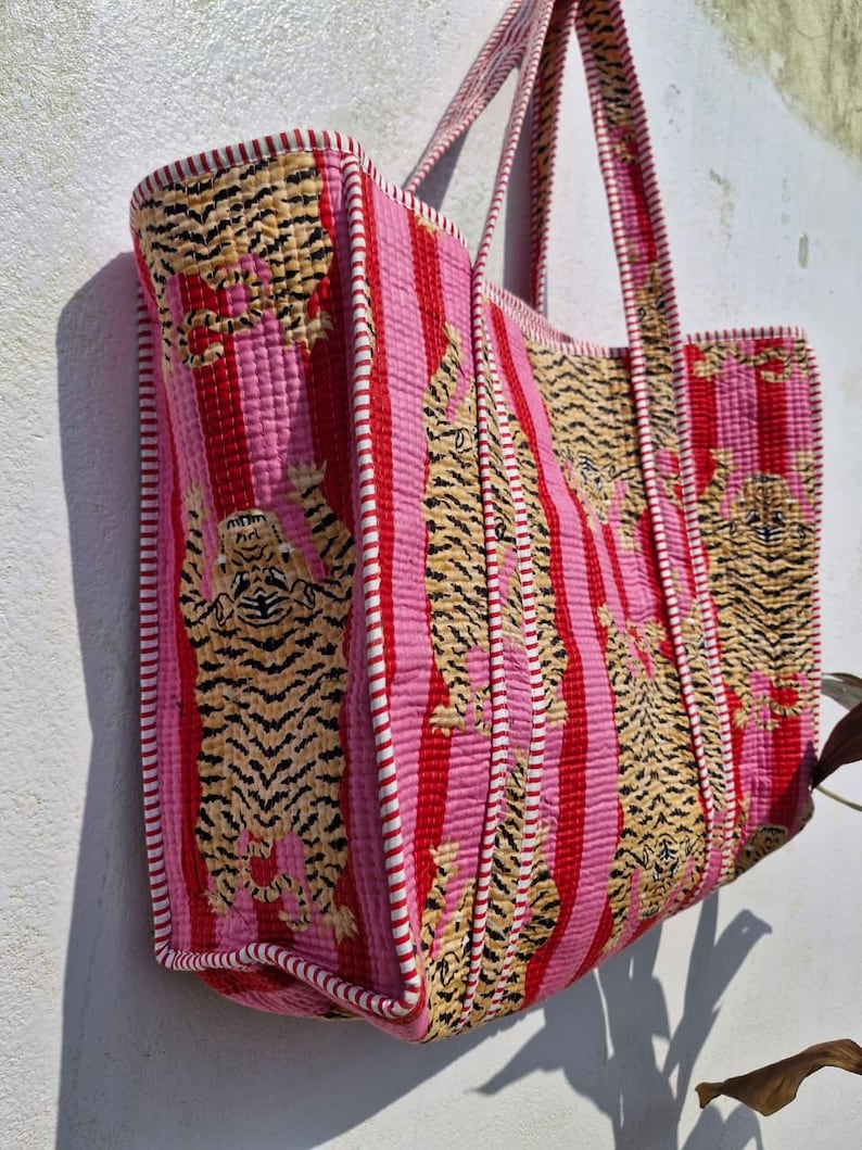 May include: A large tote bag with a quilted design. The bag features vertical stripes in pink and red, with embroidered tiger patterns. The edges are trimmed with white and red stitching, and it has matching handles.