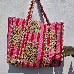May include: A large tote bag with a pink and red striped pattern. The bag features embroidered tiger designs in shades of tan and black. The handles and edges are trimmed in red.
