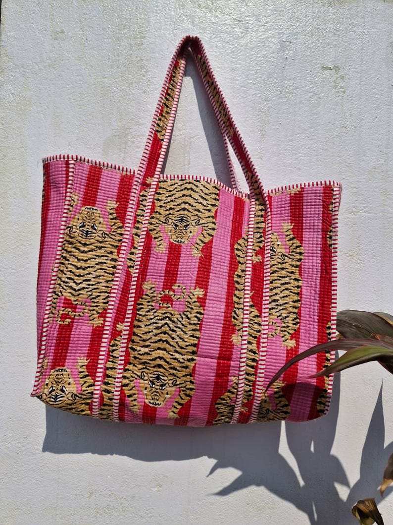 May include: A large tote bag with a pink and red striped pattern. The bag features embroidered tiger designs in shades of tan and black. The handles and edges are trimmed in red.