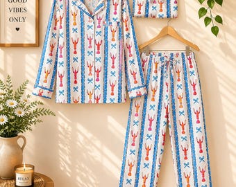 May include: A white pajama set featuring a long-sleeved button-down shirt with a chest pocket and matching pants. The fabric is decorated with a repeating pattern of red lobsters and blue bows separated by vertical blue stripes.