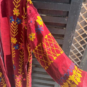 May include: A red embroidered jacket with yellow, blue, and pink floral designs. The jacket has long sleeves with yellow cross-stitch patterns and floral embroidery. The jacket has a textured, velvet-like appearance.