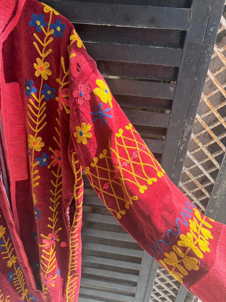 May include: A red embroidered jacket with yellow, blue, and pink floral designs. The jacket has long sleeves with yellow cross-stitch patterns and floral embroidery. The jacket has a textured, velvet-like appearance.
