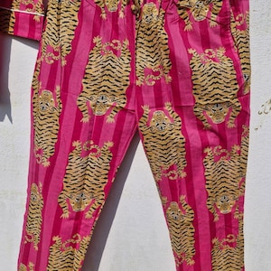 May include: Pink pajama pants with vertical stripes and a repeating tiger print in shades of tan and black. The pants have a drawstring waist and white trim along the cuffs. The pajama set is hanging on a wooden hanger.