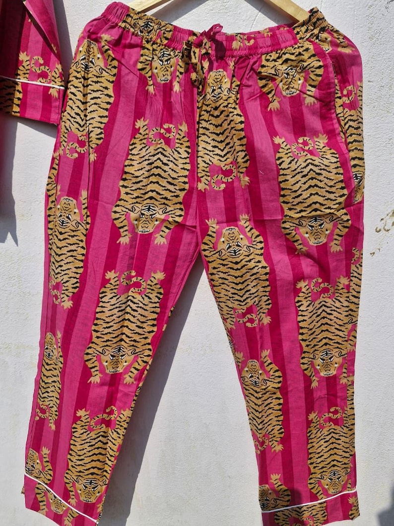 May include: Pink pajama pants with vertical stripes and a repeating tiger print in shades of tan and black. The pants have a drawstring waist and white trim along the cuffs. The pajama set is hanging on a wooden hanger.