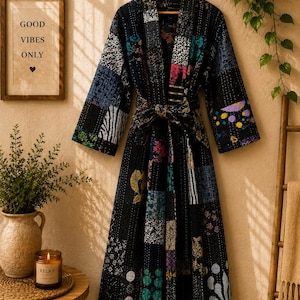 May include: A long-sleeved robe featuring a patchwork design with a black base and various colorful patterns including floral, geometric, and abstract motifs. The garment includes a matching tie belt at the waist and visible white stitching.