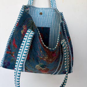 May include: A blue tote bag with floral patterns and striped lining. The bag features white and teal striped handles and trim. The interior has a pocket and a button closure. The bag's exterior has a quilted texture.