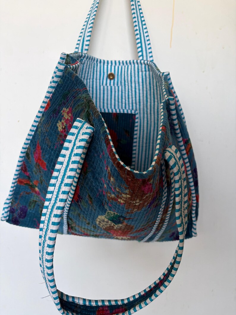 May include: A blue tote bag with floral patterns and striped lining. The bag features white and teal striped handles and trim. The interior has a pocket and a button closure. The bag's exterior has a quilted texture.