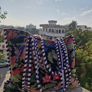May include: A large tote bag with a floral pattern in shades of red, green, and blue on a black background. The bag has thick handles with a striped design in black, white, and pink. The bag is sitting on a concrete surface.