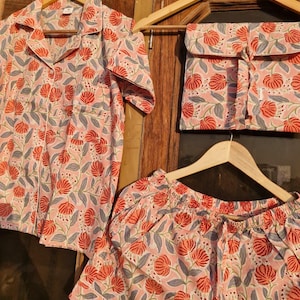 Pu&ograve; includere: Un completo pigiama con fantasia floreale nei toni del rosa, rosso e grigio. Il set comprende una camicia con bottoni, pantaloncini e un sacchetto con coulisse, il tutto appeso a grucce di legno.