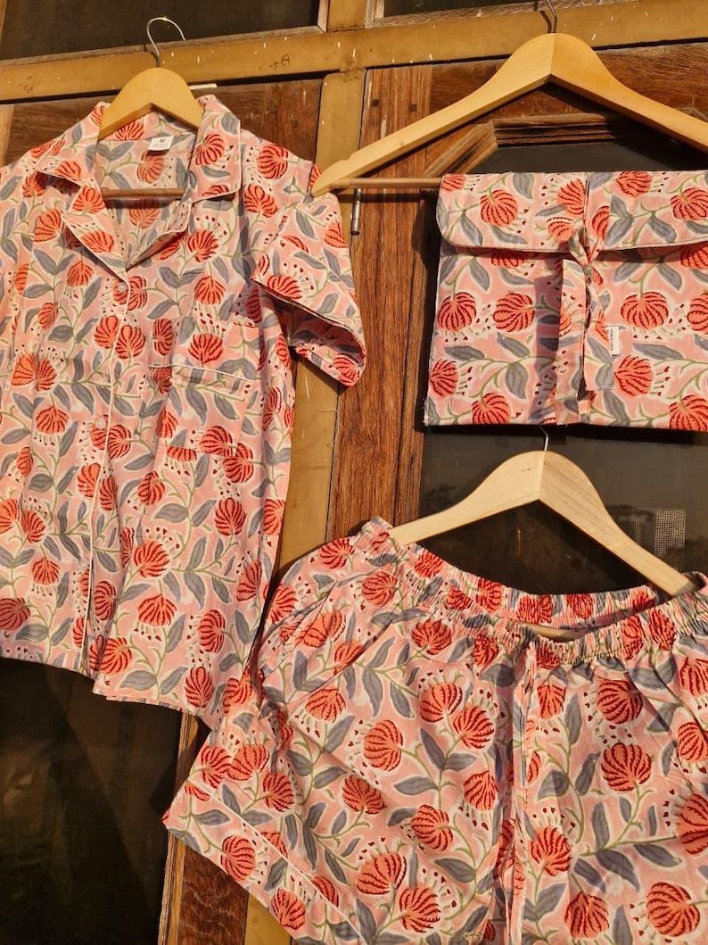 Pu&ograve; includere: Un completo pigiama con fantasia floreale nei toni del rosa, rosso e grigio. Il set comprende una camicia con bottoni, pantaloncini e un sacchetto con coulisse, il tutto appeso a grucce di legno.