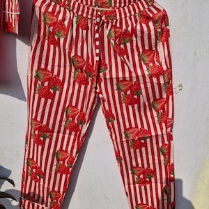 May include: Red and white striped pyjama trousers with a strawberry pattern. The trousers are hanging on a wooden hanger. The strawberries are red with green leaves. The trousers have a drawstring waist and pockets.
