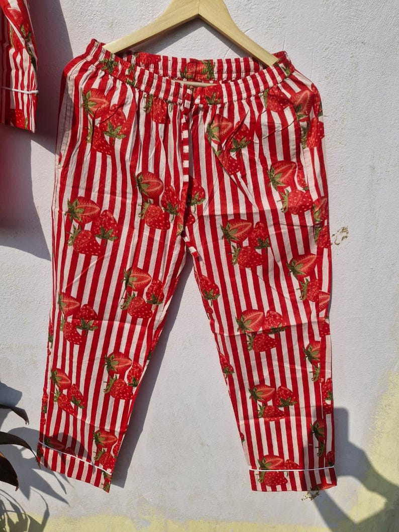 May include: Red and white striped pyjama trousers with a strawberry pattern. The trousers are hanging on a wooden hanger. The strawberries are red with green leaves. The trousers have a drawstring waist and pockets.