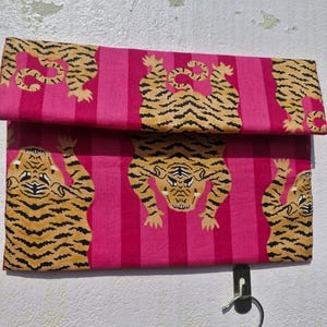 May include: A folded pink fabric with a repeating pattern of tigers in orange and black stripes. The background features vertical stripes in varying shades of pink. The fabric is hanging on a wall.
