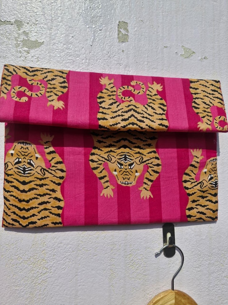 May include: A folded pink fabric with a repeating pattern of tigers in orange and black stripes. The background features vertical stripes in varying shades of pink. The fabric is hanging on a wall.