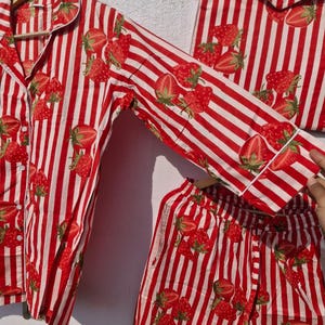 May include: Red and white striped pyjama set featuring a strawberry design. The set includes a button-down shirt, trousers, and a matching bag. The pyjamas are made from a lightweight material, designed for comfort.