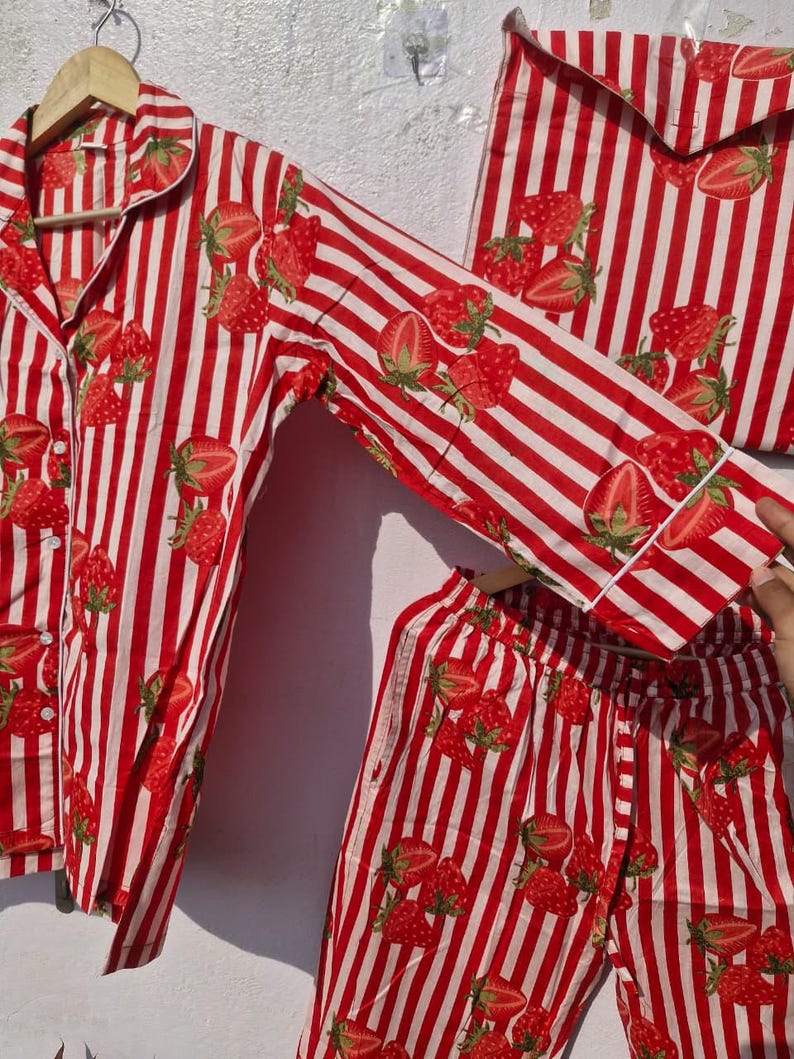 May include: Red and white striped pyjama set featuring a strawberry design. The set includes a button-down shirt, trousers, and a matching bag. The pyjamas are made from a lightweight material, designed for comfort.