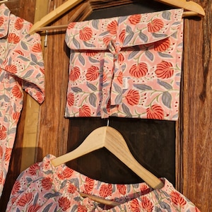 Pu&ograve; includere: Un completo pigiama rosa con un motivo floreale di fiori rossi e foglie grigie. Il set comprende un top, pantaloni e una custodia abbinata, il tutto appeso a grucce di legno contro una porta di legno.