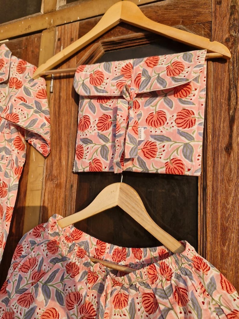 Pu&ograve; includere: Un completo pigiama rosa con un motivo floreale di fiori rossi e foglie grigie. Il set comprende un top, pantaloni e una custodia abbinata, il tutto appeso a grucce di legno contro una porta di legno.