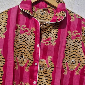 May include: Pink striped pajama top with a tiger print. The button-down shirt has white trim and is hanging on a wooden hanger. The tiger print features orange and black tigers on a pink background.