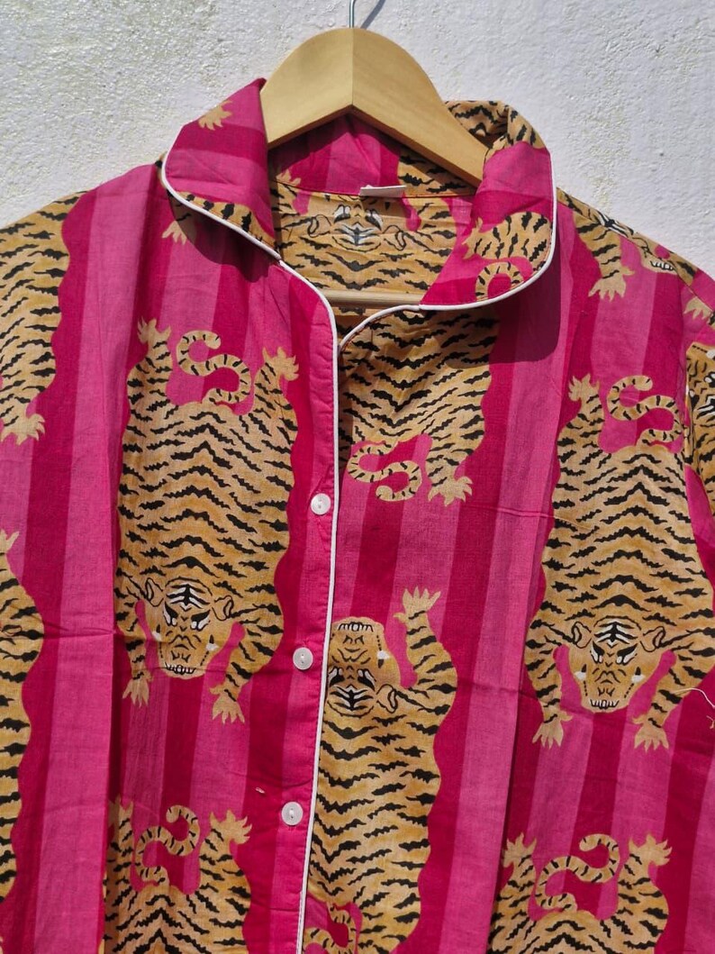 May include: Pink striped pajama top with a tiger print. The button-down shirt has white trim and is hanging on a wooden hanger. The tiger print features orange and black tigers on a pink background.
