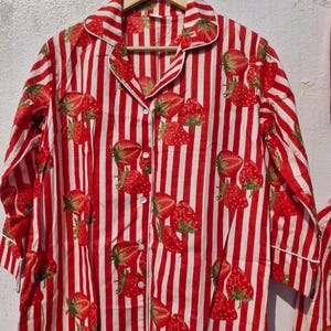 May include: A red and white striped pajama top featuring a strawberry design. The long-sleeved shirt has a button-down front, a collar, and white trim. The strawberries have green leaves and are spread across the fabric.