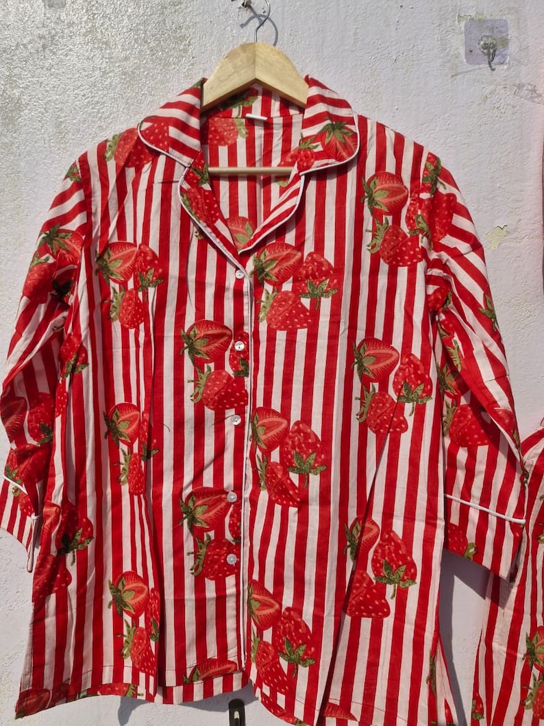 May include: A red and white striped pajama top featuring a strawberry design. The long-sleeved shirt has a button-down front, a collar, and white trim. The strawberries have green leaves and are spread across the fabric.