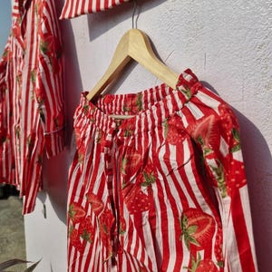 May include: A red and white striped pajama set featuring a strawberry design. The set includes a shirt, trousers, and a small pouch, all displayed on wooden hangers. The fabric appears to be lightweight.
