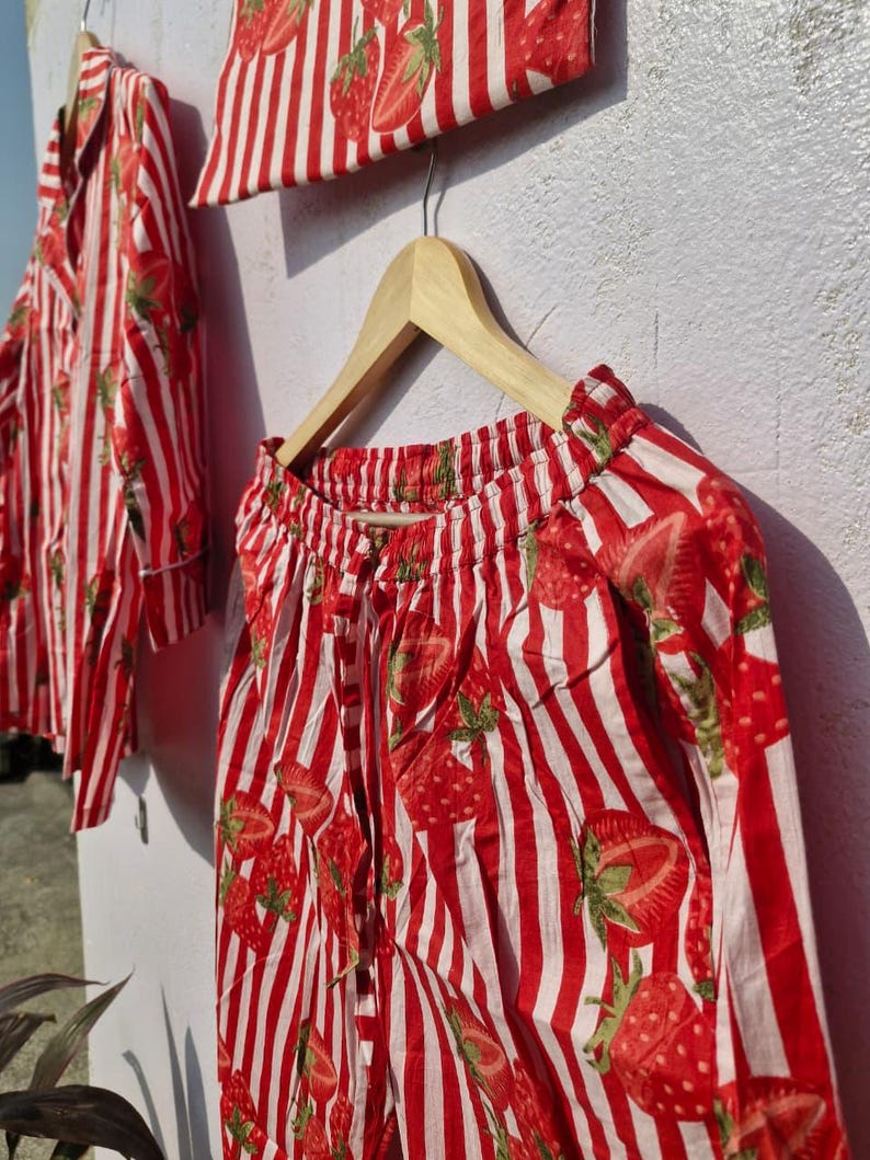 May include: A red and white striped pajama set featuring a strawberry design. The set includes a shirt, trousers, and a small pouch, all displayed on wooden hangers. The fabric appears to be lightweight.