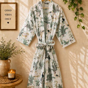 May include: A white robe featuring a repeating pattern of green palm trees, tropical foliage, and spotted leopards. The garment has long, wide sleeves, a shawl collar, and a matching fabric tie belt at the waist to secure the front closure.