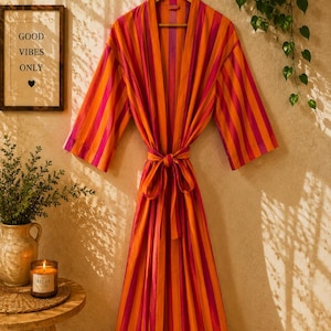 May include: A long-sleeved robe featuring vertical stripes in alternating shades of bright orange and magenta. The garment includes a matching fabric tie belt at the waist, wide sleeves, and a shawl collar, hanging from a wooden hanger.