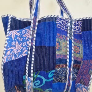 May include: A blue patchwork tote bag with a variety of fabric patterns and textures. The bag has strong handles and is trimmed with white and blue striped fabric. The design includes floral and geometric motifs.