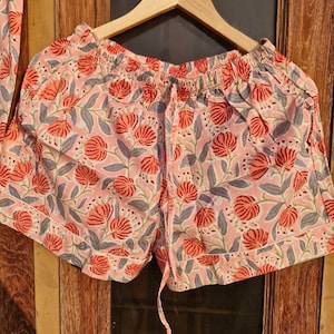 Pu&ograve; includere: Pantaloncini rosa con stampa floreale e vita con coulisse, appesi a una gruccia di legno. I pantaloncini presentano un motivo ripetuto di fiori rossi e foglie grigie su sfondo rosa chiaro. I pantaloncini fanno parte di un completo pigiama.