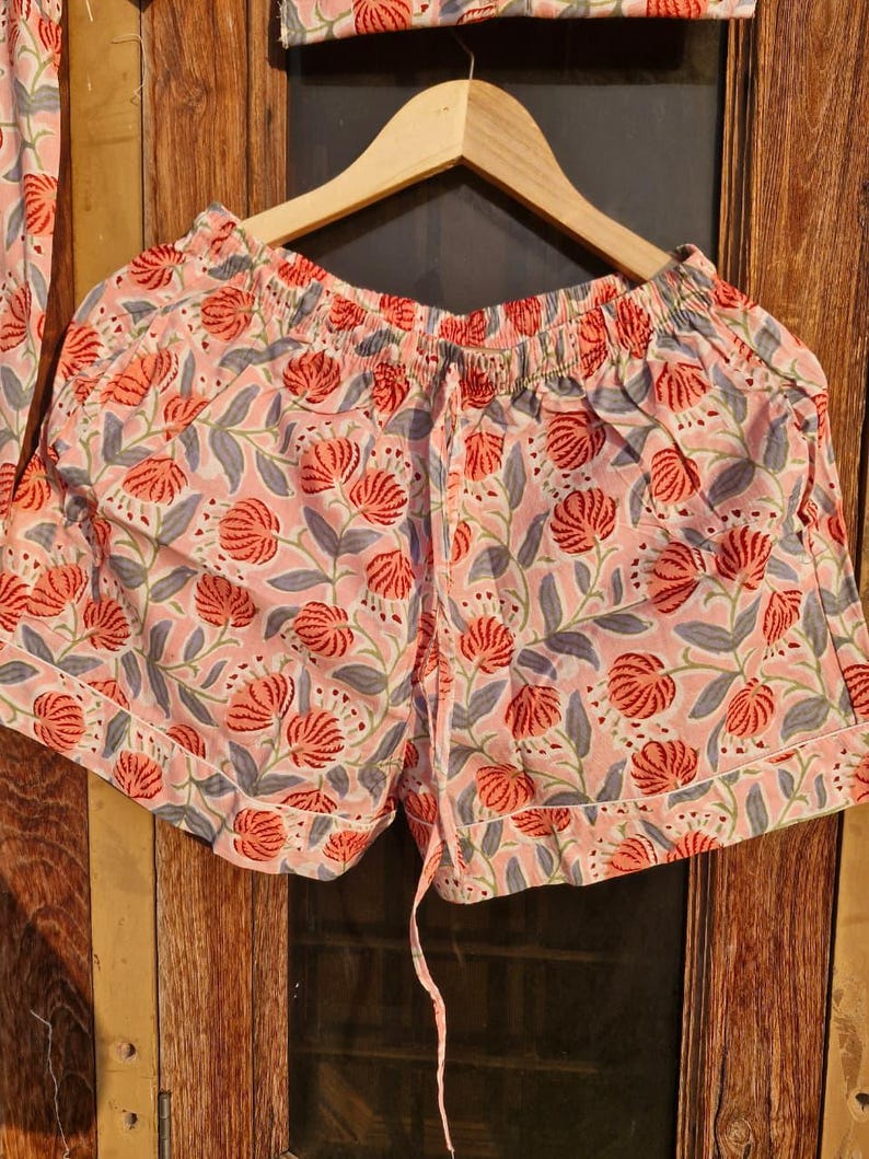 Pu&ograve; includere: Pantaloncini rosa con stampa floreale e vita con coulisse, appesi a una gruccia di legno. I pantaloncini presentano un motivo ripetuto di fiori rossi e foglie grigie su sfondo rosa chiaro. I pantaloncini fanno parte di un completo pigiama.