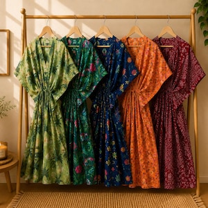 May include: Five kaftan dresses hanging on a wooden rack, each featuring a distinct floral or botanical print in shades of light green, dark green, navy blue, orange, and deep red. Each dress has a V-neckline and a cinched waist with a drawstring.