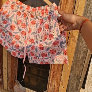 Pu&ograve; includere: Pantaloncini rosa con stampa floreale e vita con coulisse. I pantaloncini presentano un motivo di fiori rossi e foglie grigie su sfondo rosa. I pantaloncini sono appesi a una gruccia di legno.