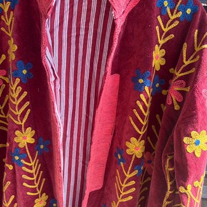 May include: A burgundy embroidered jacket with a striped interior. The jacket features floral embroidery in yellow and blue. The trim is a bright pink. The jacket is hanging on a wooden hanger.