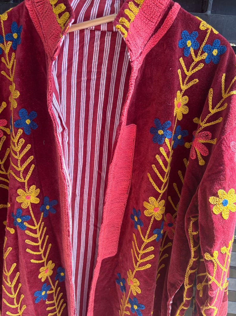 May include: A burgundy embroidered jacket with a striped interior. The jacket features floral embroidery in yellow and blue. The trim is a bright pink. The jacket is hanging on a wooden hanger.