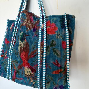 May include: A teal corduroy tote bag with a vibrant floral and bird print. The bag features a repeating pattern of red flowers, green leaves, and a hummingbird. The handles are teal with white stripes.