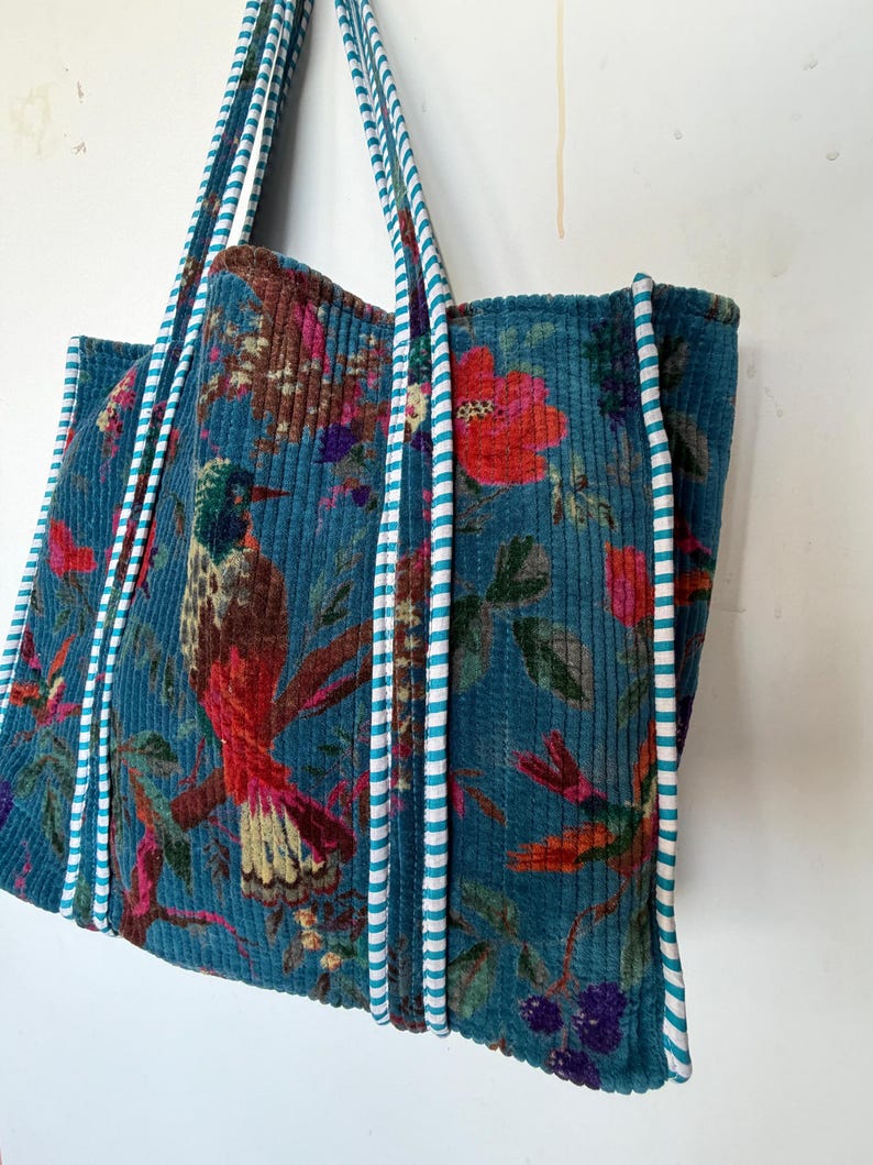 May include: A teal corduroy tote bag with a vibrant floral and bird print. The bag features a repeating pattern of red flowers, green leaves, and a hummingbird. The handles are teal with white stripes.