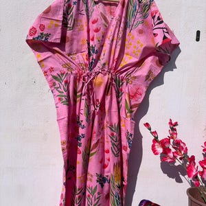 May include: A pink caftan dress with a floral print, hanging on a wooden hanger. The dress has a V-neck, drawstring waist, and a colourful pattern of flowers and leaves in pink, yellow, green, and blue. The caftan is against a white wall.
